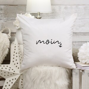 Puede incluir: Almohada blanca con texto negro que dice "moin" y un s&iacute;mbolo de ancla.