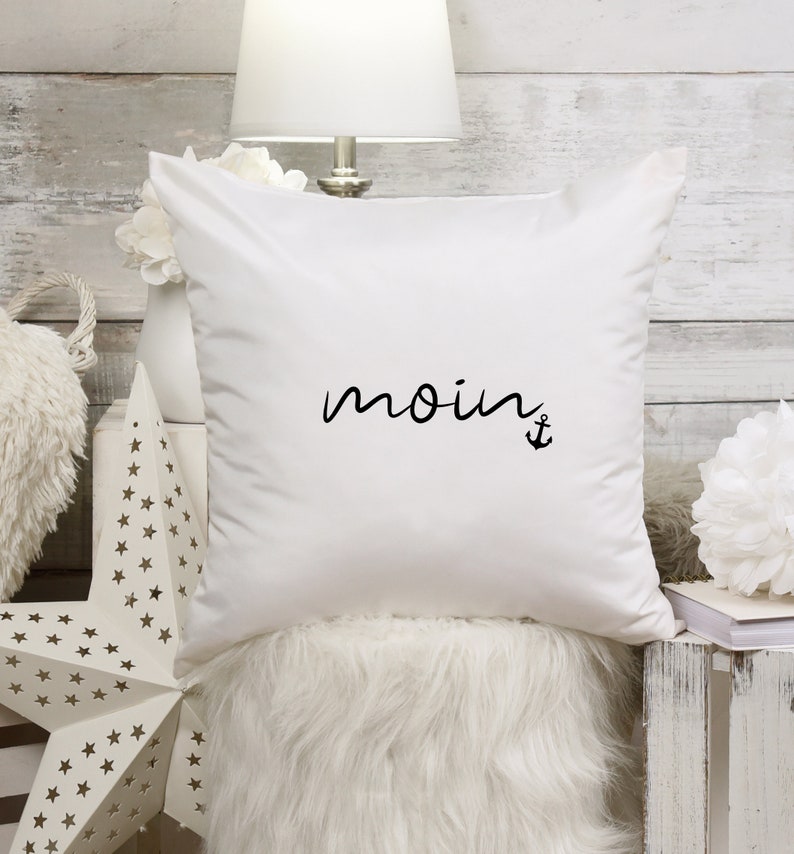 Puede incluir: Almohada blanca con texto negro que dice "moin" y un s&iacute;mbolo de ancla.