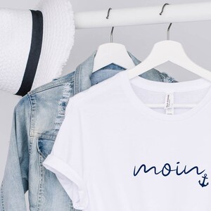 Puede incluir: Camiseta blanca con un ancla azul marino y la palabra "moin" escrita en cursiva.