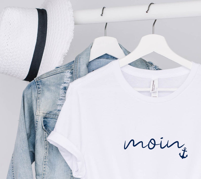 Puede incluir: Camiseta blanca con un ancla azul marino y la palabra "moin" escrita en cursiva.