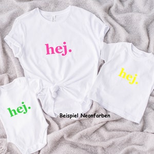 Puede incluir: Tres camisetas blancas con la palabra "hej" impresa en ellas en diferentes colores ne&oacute;n: rosa, amarillo y verde. Las camisetas est&aacute;n dispuestas en forma de pir&aacute;mide sobre una superficie gris y peluda. El texto "Beispiel Neonfarben" es visible debajo de las camisetas.