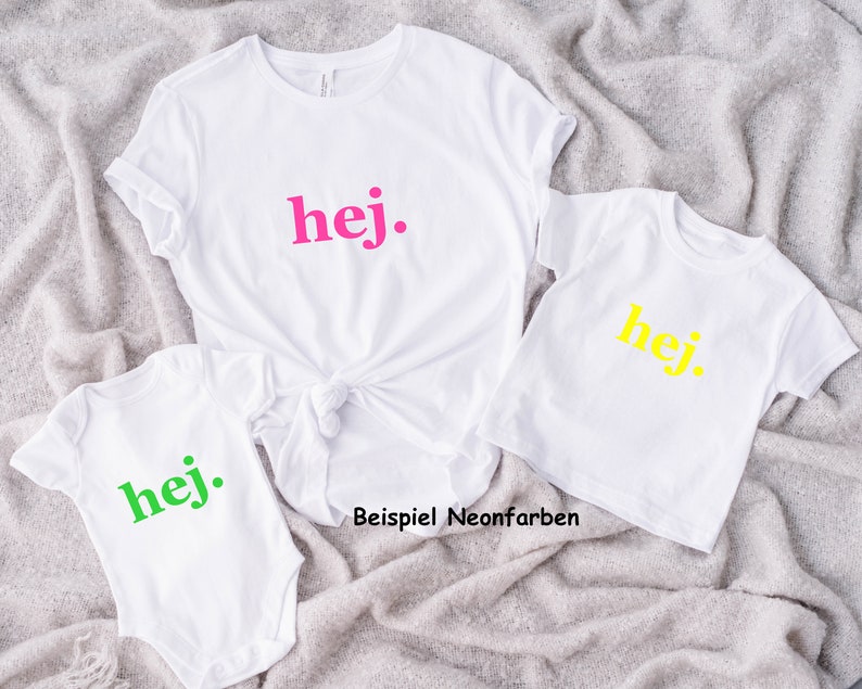 Puede incluir: Tres camisetas blancas con la palabra "hej" impresa en ellas en diferentes colores ne&oacute;n: rosa, amarillo y verde. Las camisetas est&aacute;n dispuestas en forma de pir&aacute;mide sobre una superficie gris y peluda. El texto "Beispiel Neonfarben" es visible debajo de las camisetas.