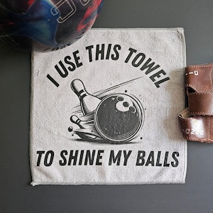 May include: A square grey microfiber towel featuring a black graphic of a bowling ball and pin with the text I USE THIS TOWEL TO SHINE MY BALLS printed in black. The towel is positioned next to a multi-colored bowling ball and a brown leather wrist support.