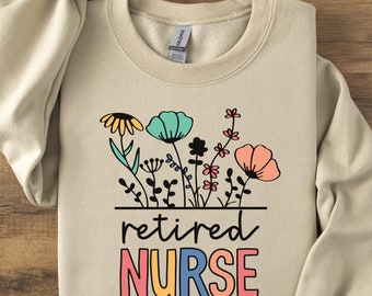 May include: A beige crewneck sweatshirt featuring a graphic of colorful wildflowers above the black text "retired" and the multi-colored block letters "NURSE". The design includes yellow, teal, pink, and black floral elements on the front.