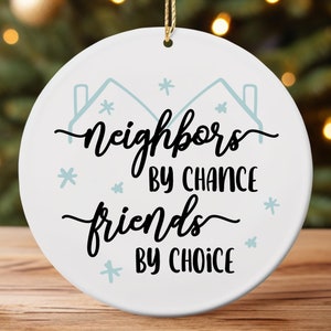 May include: White ceramic Christmas ornament with the text "neighbors by chance friends by choice" in black script. The ornament has light blue house and snowflake designs. A gold string is attached for hanging.