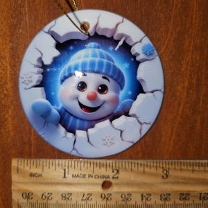 May include: A round Christmas ornament featuring a smiling snowman wearing a blue hat and scarf, emerging from a cracked white surface. The snowman has a red nose and black eyes. The ornament is approximately 3 inches in diameter.