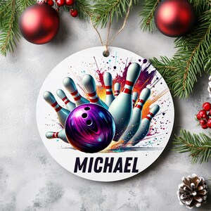 May include: A white, round Christmas ornament featuring a bowling theme. The design includes a purple bowling ball, white bowling pins with red stripes, and the name "MICHAEL" in bold black letters. The background has colorful paint splatters.