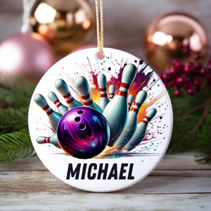 May include: A round, white bowling-themed ornament with a vibrant design. It features a purple and black bowling ball, white bowling pins with red stripes, and the name "MICHAEL" in bold black letters. The ornament hangs from a gold chain.