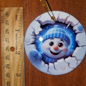 May include: Round Christmas ornament featuring a smiling snowman wearing a blue hat and scarf. The ornament is approximately 3 inches in diameter and has a gold string for hanging. The snowman is emerging from a broken ice effect.