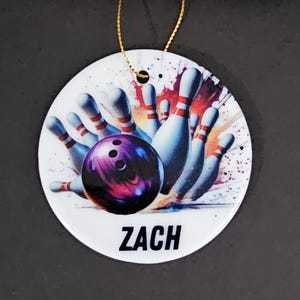 May include: A white, round ornament with a bowling theme. The design features a purple bowling ball striking pins, with the name "ZACH" in black text. A gold string is attached for hanging.