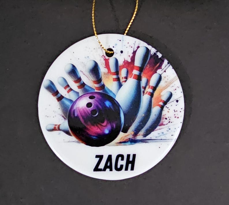 May include: A white, round ornament with a bowling theme. The design features a purple bowling ball striking pins, with the name "ZACH" in black text. A gold string is attached for hanging.