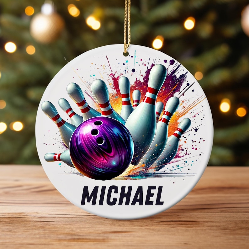 May include: A white ceramic ornament featuring a bowling scene with a purple bowling ball and white pins with red stripes. The word "MICHAEL" is printed in black at the bottom. A gold string is attached for hanging.