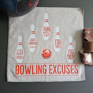 May include: A square gray towel featuring five white bowling pins and a red bowling ball. Each pin has red text: SLICK APPROACH, USER ERROR I GUESS, THUMB STUCK, TOO TIRED, and TOO OILY. The bottom text reads BOWLING EXCUSES in large red letters.