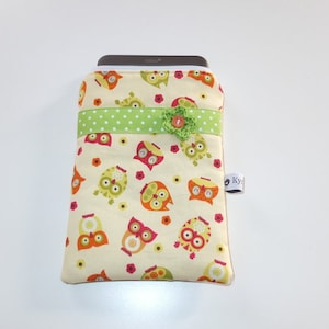 Könnte beinhalten: Eine cremefarbene Stofftasche mit einem Muster aus bunten Eulen ist mit einem grün-weiß gepunkteten Band und einer grünen Blumenverzierung geschmückt. Die Tasche hat ein weißes Etikett mit dem Text "Ky" darauf.