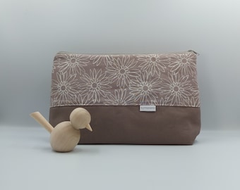 Op de afbeelding: Een rechthoekige, taupekleurige etui met ritssluiting en een bloemenpatroon op de bovenste helft. De bloemen zijn wit en de onderste helft is effen, donkerder bruin. Een klein houten vogelbeeldje staat op de voorgrond. De etui heeft de tekst "kymarami".