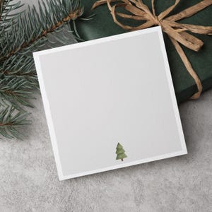 Könnte beinhalten: Eine quadratische, cremefarbene Karte mit einer Aquarellillustration eines grünen Weihnachtsbaums in der unteren rechten Ecke. Die Karte ist von einem dünnen, weißen Rahmen umgeben. Sie liegt neben einer grünen Geschenkbox mit braunem Band und Tannenzweigen.