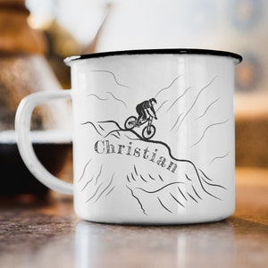 Könnte beinhalten: Eine weiße Emailletasse mit schwarzem Rand. Die Tasse zeigt eine Schwarzweißillustration eines Mountainbikers, der einen Bergpfad hinunterfährt. Das Wort "Christian" ist in einer stilisierten Schrift unter dem Biker geschrieben.