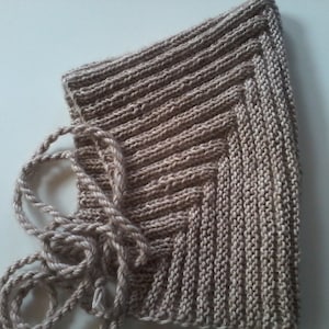 Puede incluir: Una capucha de punto beige con un cordón largo anudado. La capucha está hecha con un patrón de punto simple y tiene una superficie ligeramente texturizada.