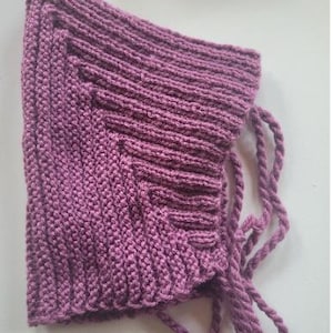 Pode incluir: Um gorro de malha na cor malva com uma textura canelada distinta e uma silhueta pontiaguda. A peça inclui longos cordões de fio torcido presos nas laterais para fixação, exibindo um padrão de malha consistente em toda a sua extensão.