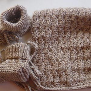 Op de afbeelding: Een set gebreide babyschoentjes en een bijpassend dekentje in een lichtbruine kleur. De schoentjes hebben een simpel ontwerp en het dekentje heeft een textuurpatroon.