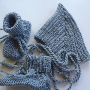 Op de afbeelding: Een set van grijze gebreide babyschoentjes en een bijpassende muts met een puntige top. De schoentjes hebben veters en de muts heeft een lang, dun koord.