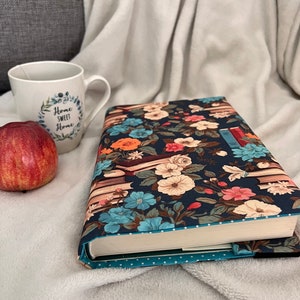 Könnte beinhalten: Ein Buchcover mit einem Blumenmuster mit blauen, rosa und orangefarbenen Blumen auf dunkelblauem Hintergrund. Das Cover hat ein weißes Punkte-Muster am unteren Rand. Der Text "Home Sweet Home" ist auf einer weißen Tasse im Hintergrund zu sehen.