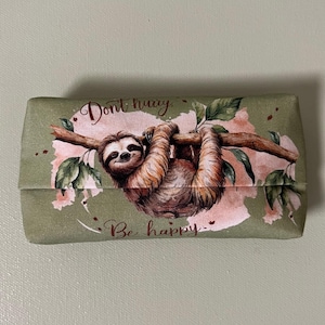 Op de afbeelding: Groene stoffen tissueboxhoes met een luiaard die aan een tak hangt. De luiaard is bruin en heeft een roze neus. De tekst op de hoes luidt "Haast je niet, Wees gelukkig."
