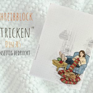 Könnte beinhalten: A5-Notizblock mit dem Text "SCHREIBBLOCK STRICKEN" und einer Illustration einer strickenden Person. Die Illustration zeigt eine Person, die einen rot-grün gestreiften Schal strickt, während eine Katze in der Nähe schläft. Einseitig bedruckt.