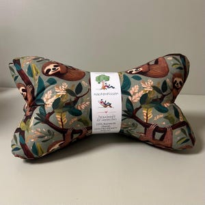 May include: A green and brown neck pillow with a sloth pattern. The pillow is made of 100% cotton and has a 100% polyester filling. The pillow is labelled "Nackenkissen" which is German for "neck pillow".