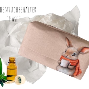 Op de afbeelding: Een lichtroze tissuebox met een illustratie van een konijn met een oranje sjaal en een witte mok. Daarnaast liggen witte tissues, een klein flesje olie en aloë vera bladeren. De tekst op de doos luidt "TASCHENTUCHBEHÄLTER 'HASE'".