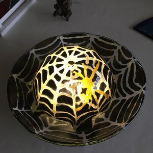 May include: A black and white glass bowl with a spider web design and a yellow spider inside. The bowl is lit from within, creating a glowing effect.