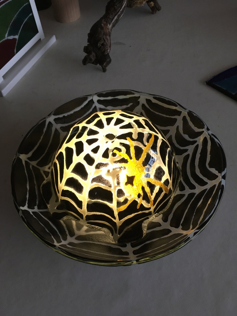 May include: A black and white glass bowl with a spider web design and a yellow spider inside. The bowl is lit from within, creating a glowing effect.