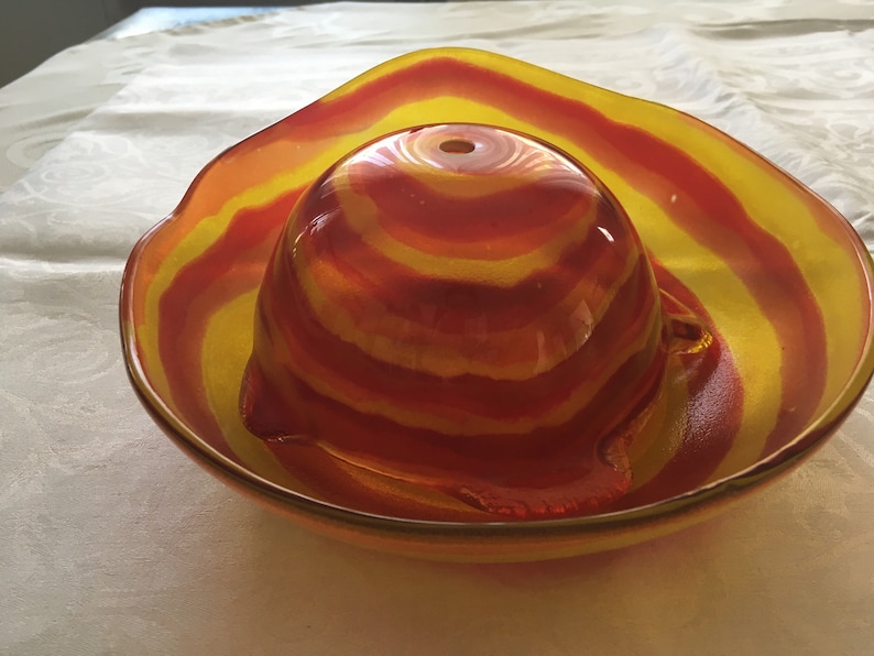 Op de afbeelding: Een rode en gele gestreepte glazen kom met een kleinere, bijpassende kom erin. De kommen zijn gemaakt van helder glas met een draaiend patroon van rood en geel.
