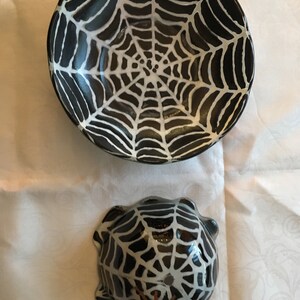 May include: Two black ceramic bowls with white spiderweb designs. The larger bowl has a simple spiderweb design, while the smaller bowl has a more detailed spiderweb design with a brown spider in the center.