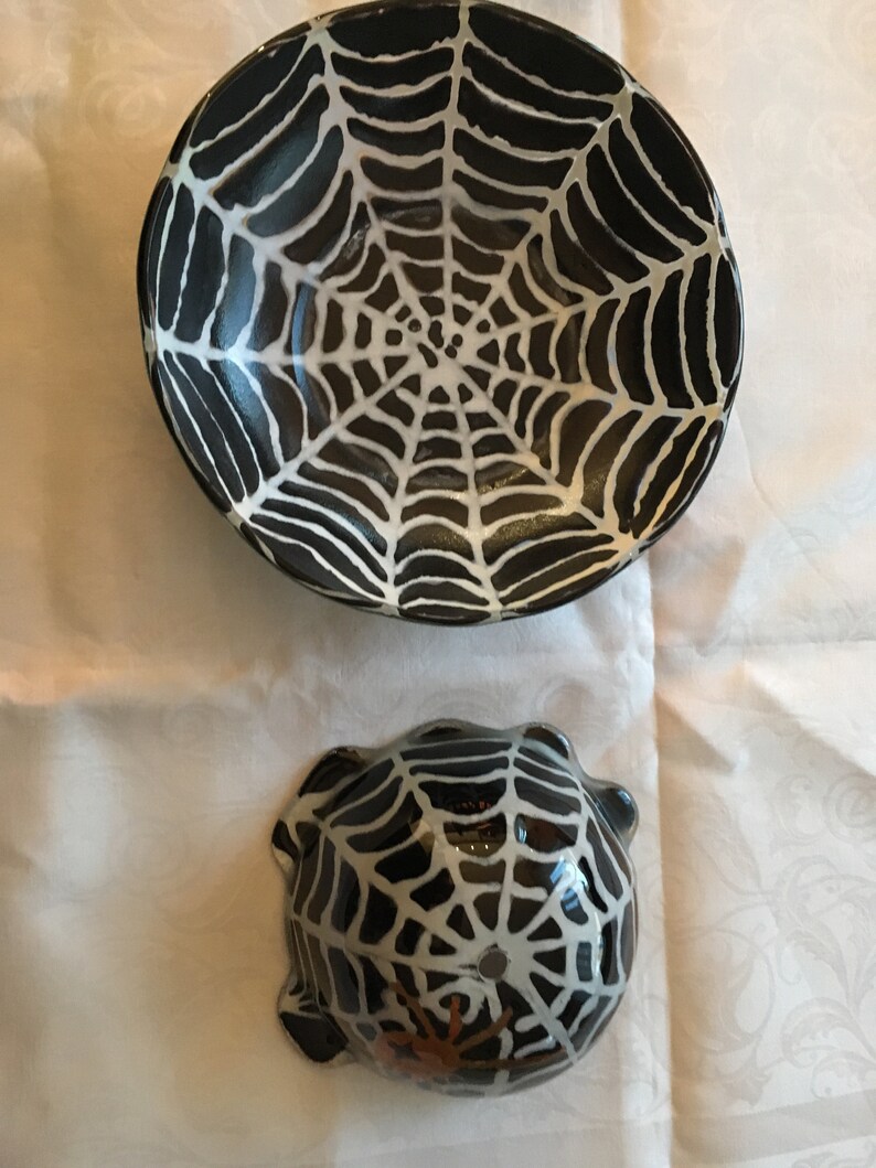 May include: Two black ceramic bowls with white spiderweb designs. The larger bowl has a simple spiderweb design, while the smaller bowl has a more detailed spiderweb design with a brown spider in the center.