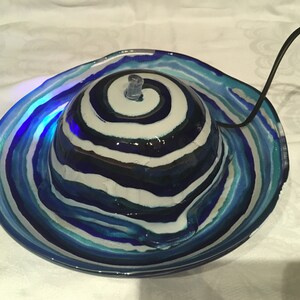 Op de afbeelding: Een blauwe en witte glazen waterfontein met een swirl-patroon. De fontein heeft een kleine pomp en een snoer.