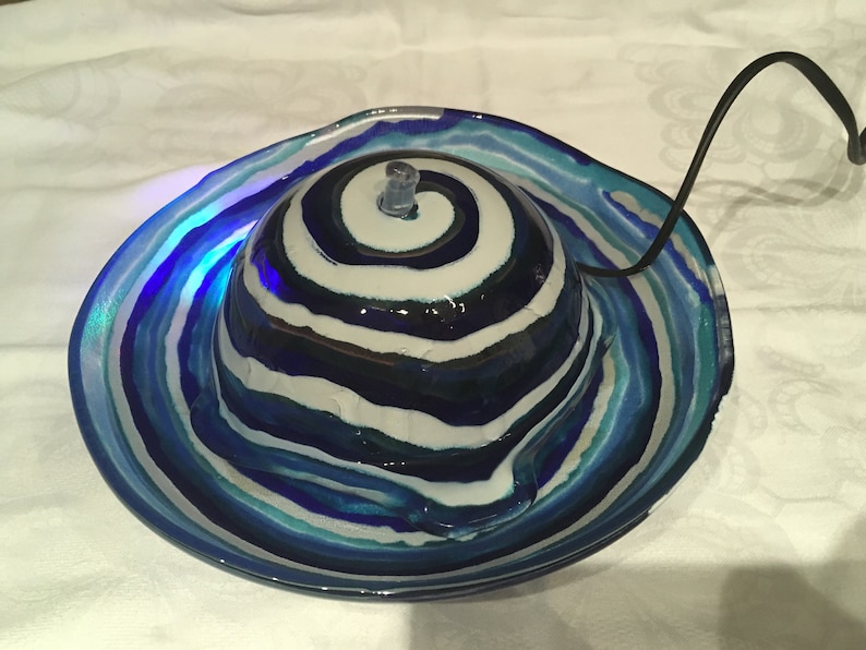 Op de afbeelding: Een blauwe en witte glazen waterfontein met een swirl-patroon. De fontein heeft een kleine pomp en een snoer.