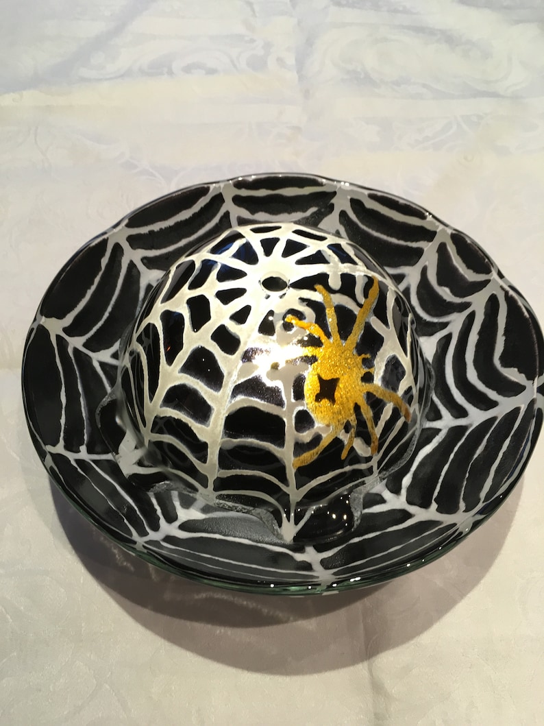 May include: A black glass bowl with a white spiderweb design and a gold spider in the center. The bowl is perfect for serving snacks or desserts.