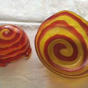 Op de afbeelding: Twee glazen kommen met een rood en geel swirl-patroon. De kommen zijn gemaakt van helder glas en hebben een glanzende afwerking.