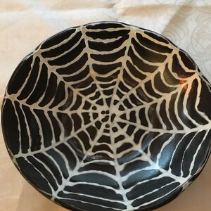 May include: A black ceramic bowl with a white spiderweb design. The bowl is perfect for serving Halloween treats or holding spooky decorations.
