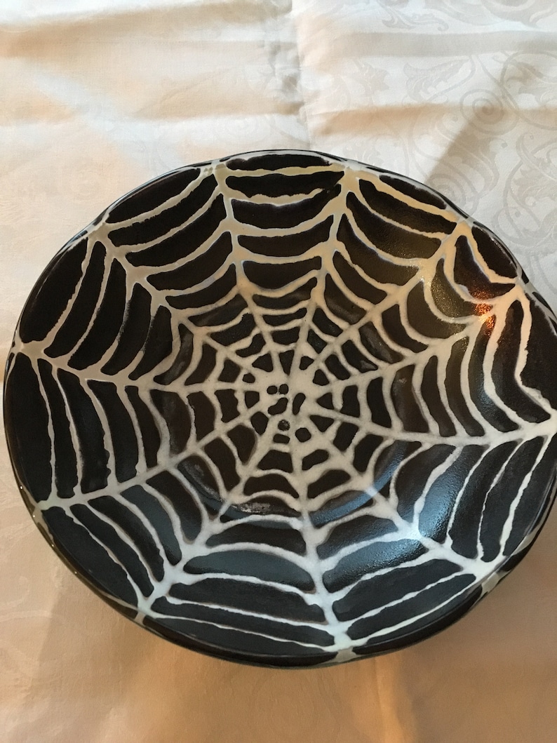 May include: A black ceramic bowl with a white spiderweb design. The bowl is perfect for serving Halloween treats or holding spooky decorations.