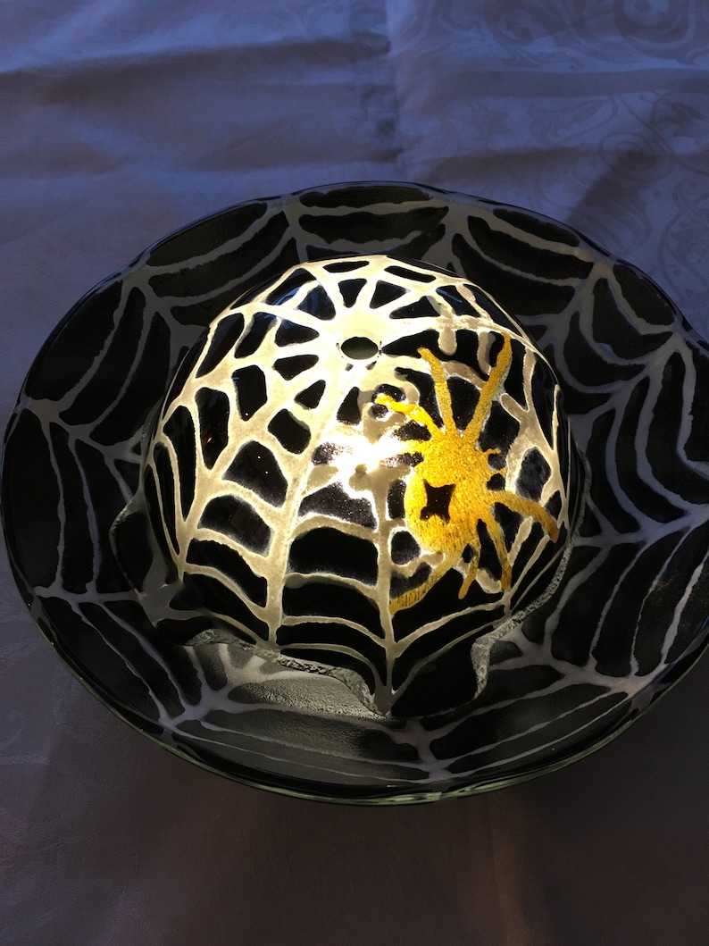 May include: A black and white glass dome with a spider web design and a gold spider inside. The dome is lit from within, creating a glowing effect.