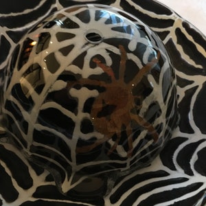 May include: A black and white ceramic dish with a spider web design. A clear glass dome sits on top of the dish, with a brown spider inside.