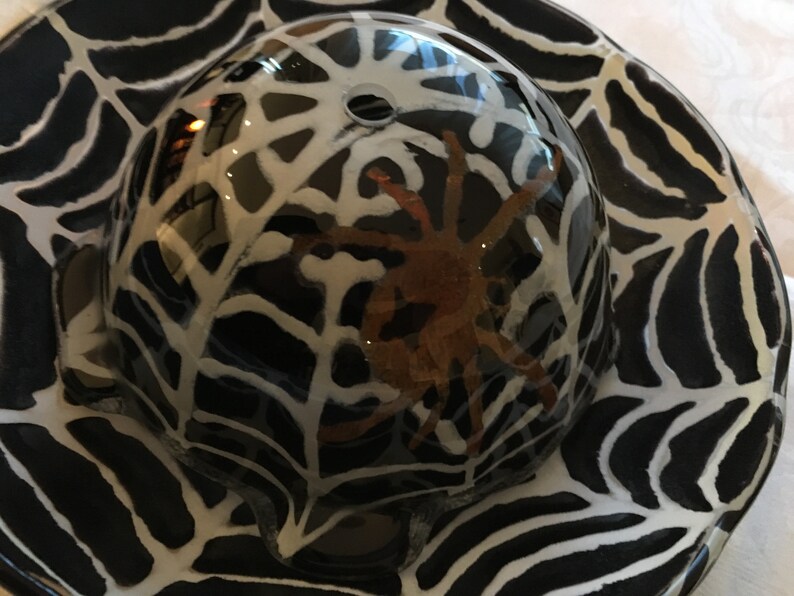 May include: A black and white ceramic dish with a spider web design. A clear glass dome sits on top of the dish, with a brown spider inside.