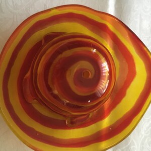 Op de afbeelding: Een glazen kom met een rood en geel swirl-patroon. De kom is rond en heeft een klein gaatje in het midden.