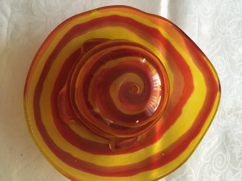Op de afbeelding: Een glazen kom met een rood en geel swirl-patroon. De kom is rond en heeft een klein gaatje in het midden.