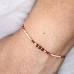 Könnte beinhalten: Ein zartes Perlenarmband mit einem sich wiederholenden Muster aus kleinen cremefarbenen, dunkelroten und goldfarbenen Metallperlen. Das Design enthält drei größere, facettierte dunkelrote Perlen, die einen dezenten Blickfang bilden.