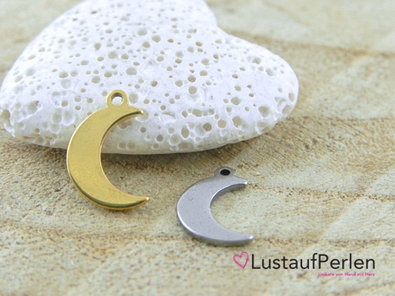 May include: Two crescent moon charms, one gold and one silver, on a white heart-shaped stone. The charms have a hole at the top for attaching to jewelry.