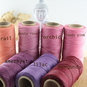 Könnte beinhalten: Spulen mit gewachstem Baumwollgarn in verschiedenen Schattierungen von Rosa, Lila und Rot. Die Farben sind mit "Koralle", "Staubrose", "Orchidee", "Bonbonrosa", "Amethyst", "Lila" und "Burgunder" beschriftet.
