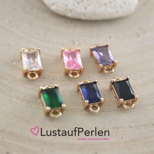 Könnte beinhalten: Sechs rechteckige Edelsteinanhänger in Goldfassungen. Die Edelsteine sind klar, rosa, lila, grün, blau und schwarz. Jeder Anhänger hat unten eine kleine Schlaufe. Der Text "LustaufPerlen" ist sichtbar.
